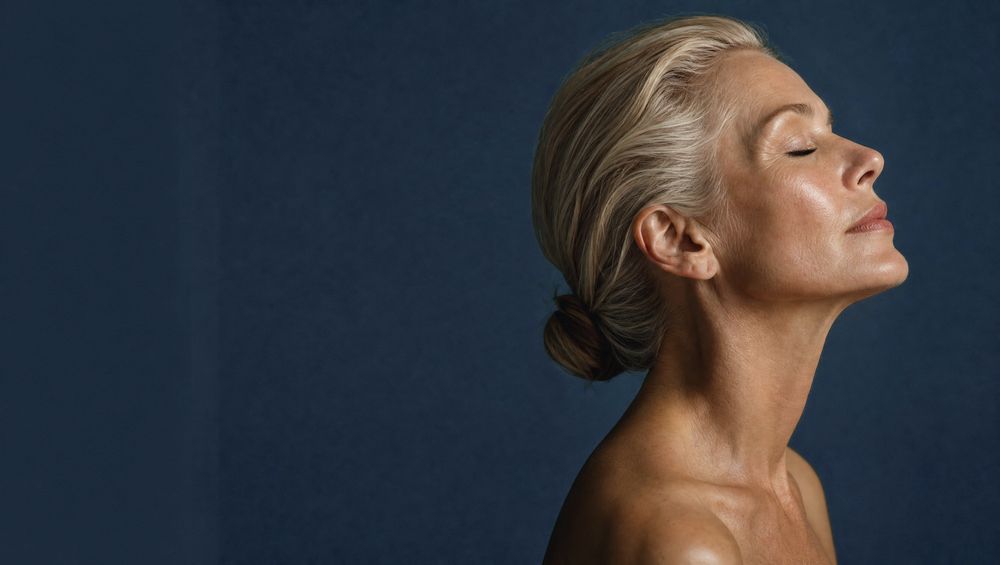 Woman in her fifties in profile with glowing skin against a deep blue backdrop
